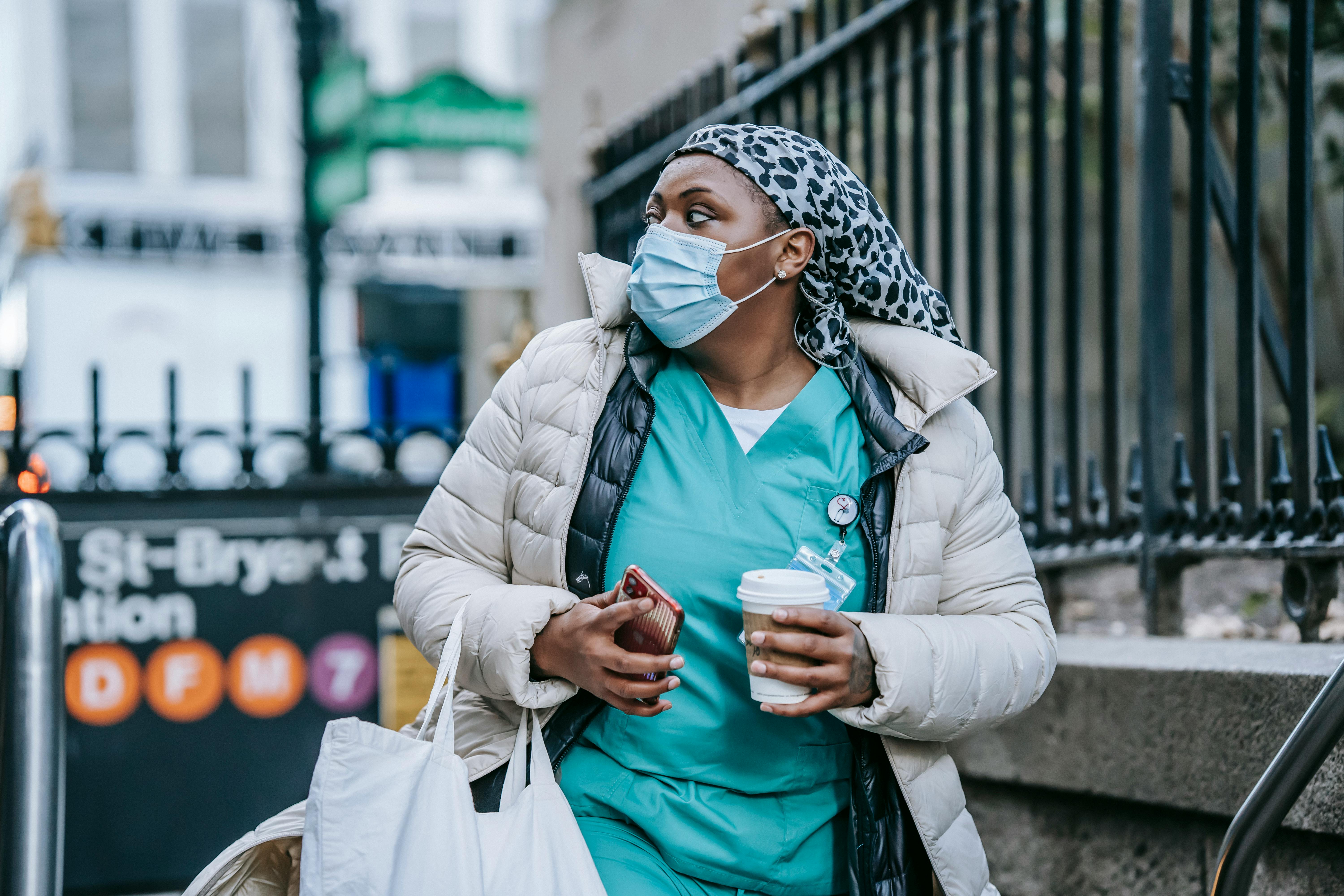 A masked healthcare worker in scrubs carrying coffee while commuting outdoors.