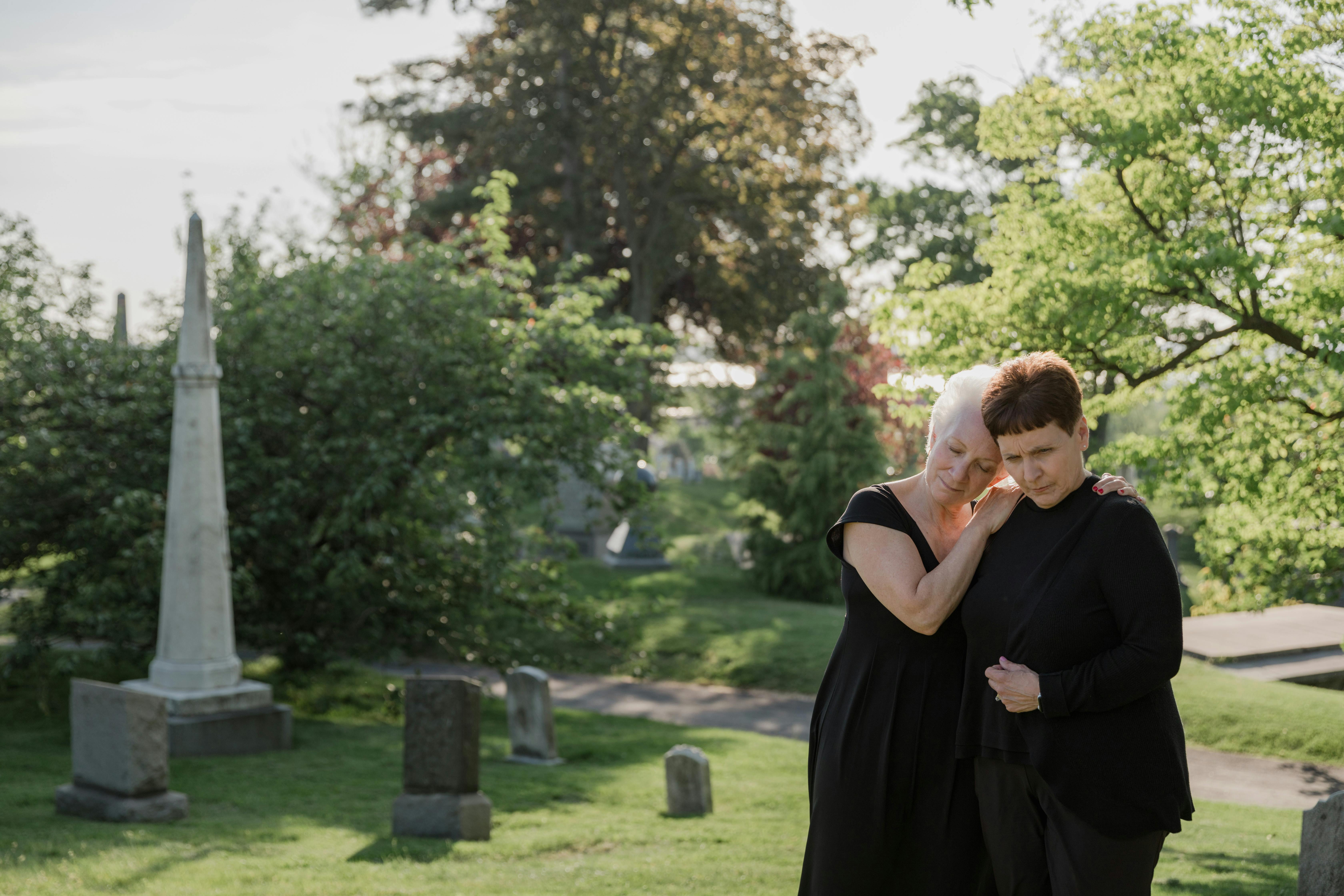 Two women in black clothing mourn in a peaceful cemetery setting.