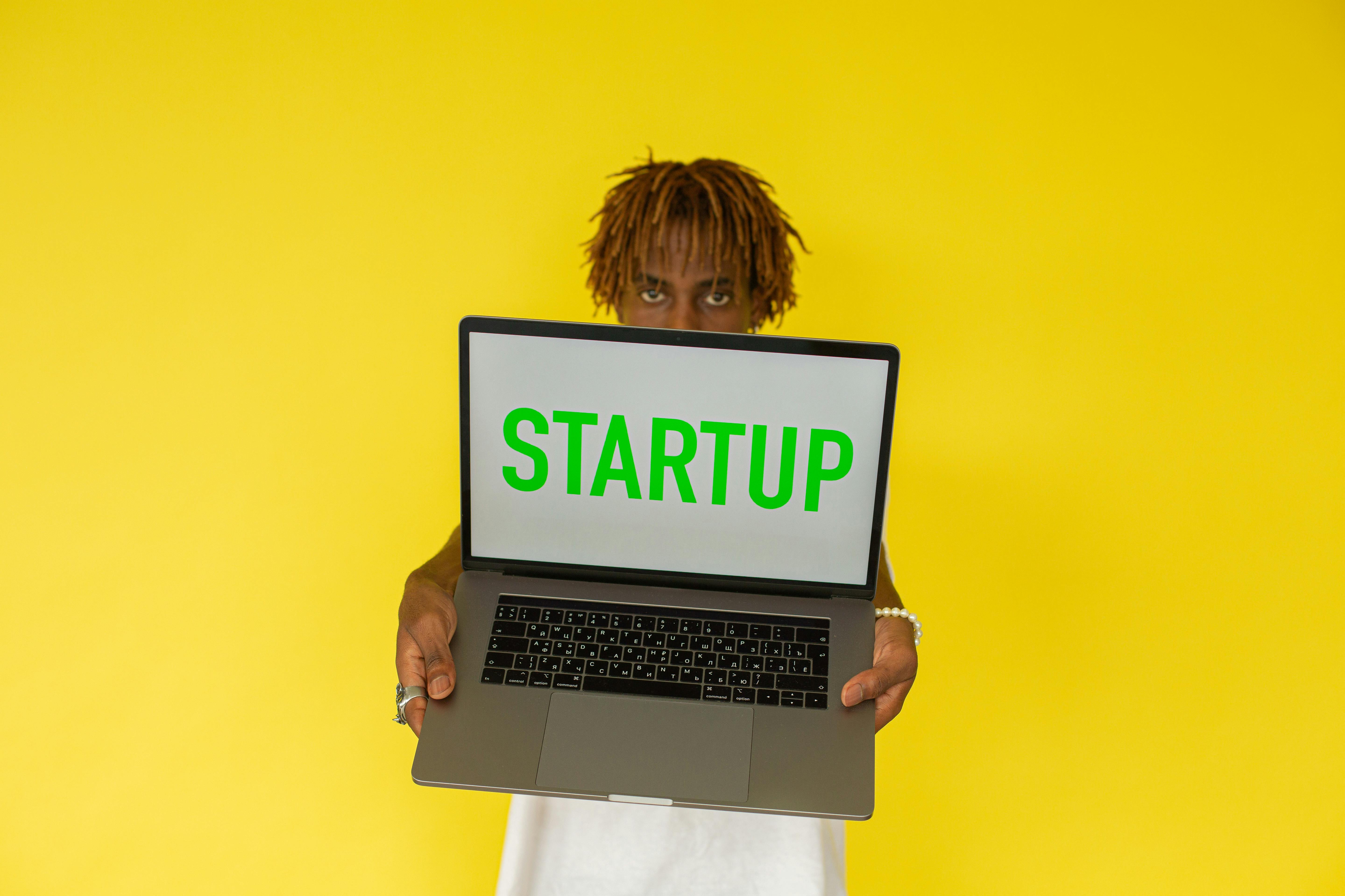 A young black man holds a laptop displaying 'Startup' against a vibrant yellow background.